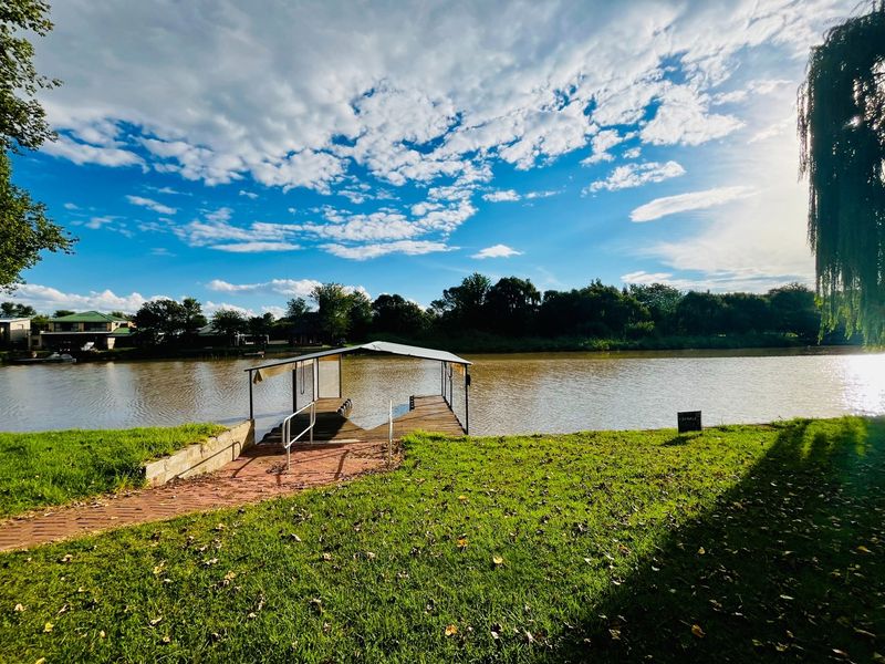 A scenic view of the riverfront featuring a private boat jetty with a covered slip, brick pathway, and landscaped lawn.