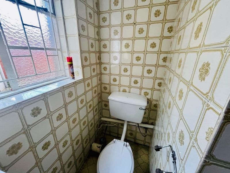 A compact, dated bathroom featuring original patterned ceramic wall tiles, a standard toilet, and a window with security bars.