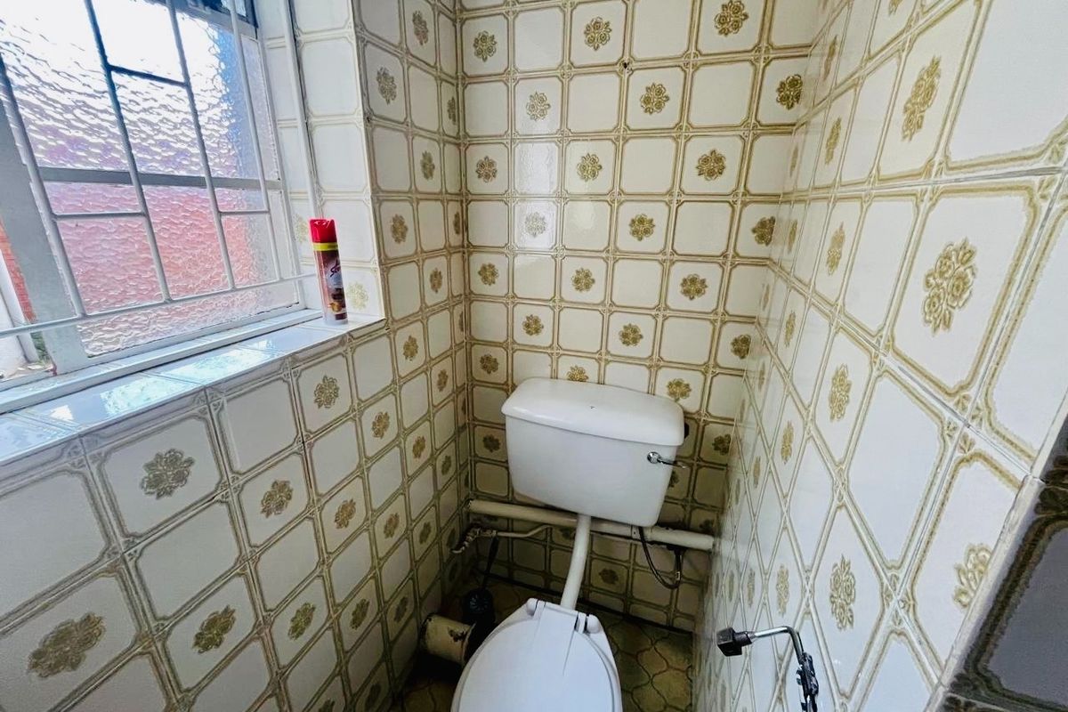 A compact, dated bathroom featuring original patterned ceramic wall tiles, a standard toilet, and a window with security bars.
