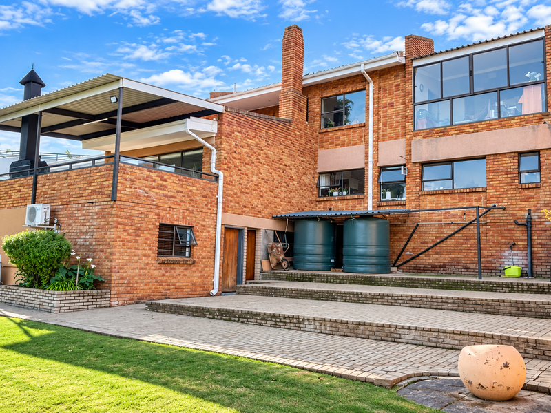 Rear view of a multi-level brick house featuring a covered patio, outdoor fireplace, paved terrace, and integrated water storage tanks in a landscaped garden.