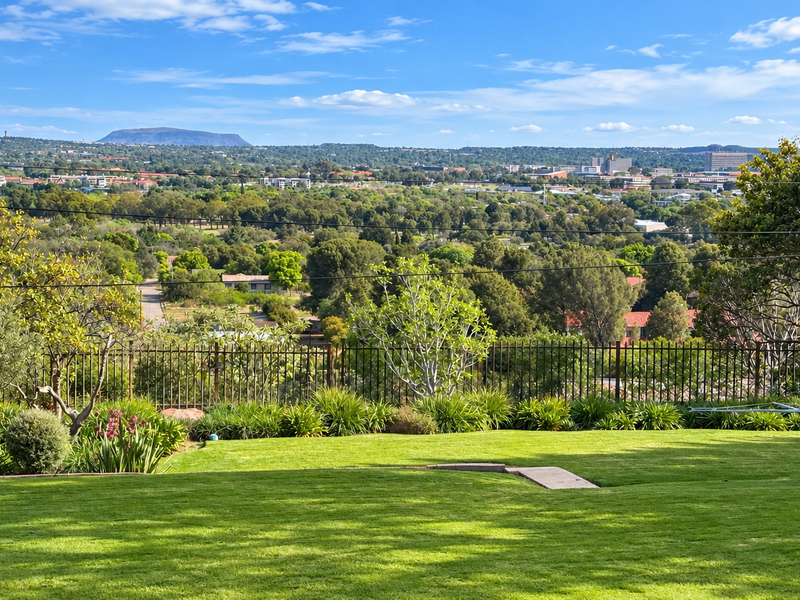 A scenic elevated view overlooking a lush green garden, mature trees, and a sprawling suburban landscape under a clear blue sky.
