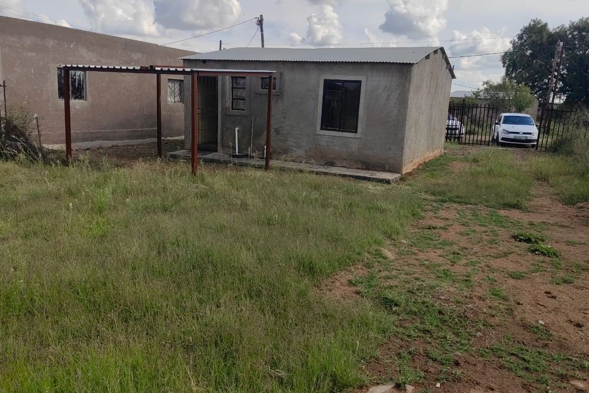 A detached residential structure featuring an unfinished plaster exterior, corrugated metal roof, and a basic metal patio frame set within a large grassed yard.