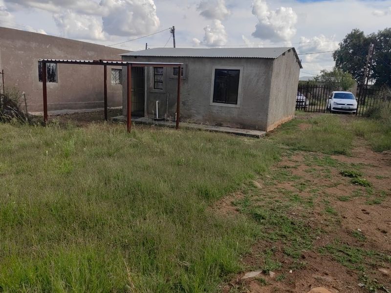 A detached residential structure featuring an unfinished plaster exterior, corrugated metal roof, and a basic metal patio frame set within a large grassed yard.