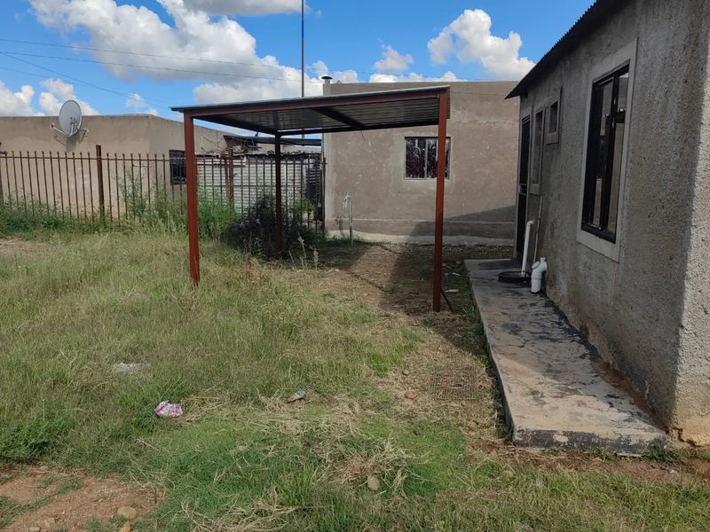 Rear exterior view of the property featuring a metal carport structure, concrete walkway, and perimeter steel fencing with an unlandscaped garden area.