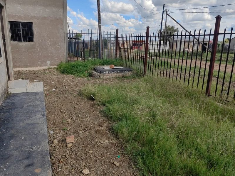Rear exterior area featuring metal security fencing, an inspection cover, and a window with security bars, set against an unmaintained garden space.