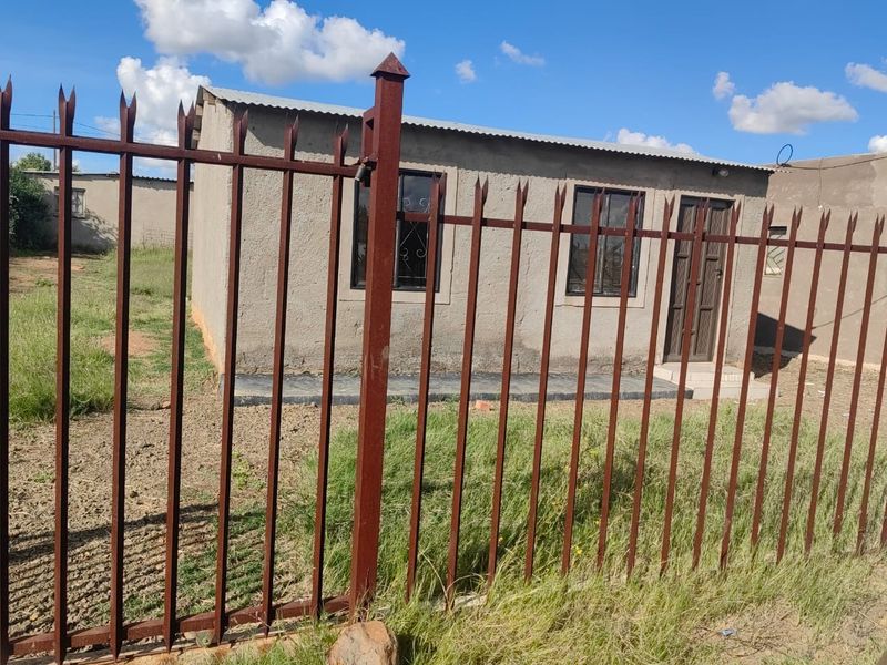 A modest single-story house featuring a corrugated metal roof, security fencing, and security bars on the windows, set on a basic plot.