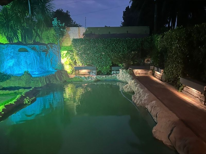A landscaped pool area featuring a rock waterfall, integrated lighting, and wooden decking, creating an atmospheric outdoor entertainment space.