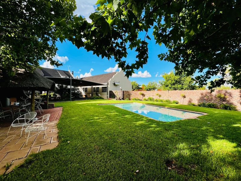 A well-maintained garden featuring a swimming pool, a thatched lapa, and a paved patio area, set against a brick boundary wall.