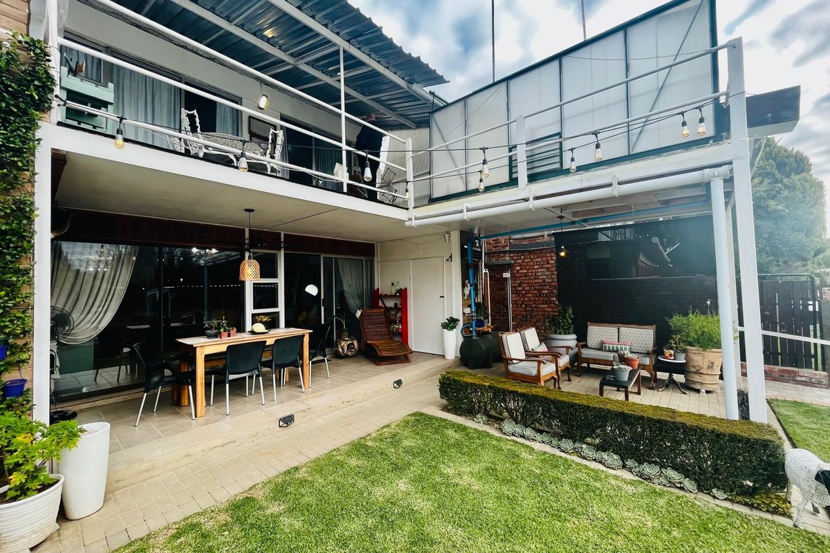 A spacious rear exterior featuring a covered patio area, an upper-level balcony, and a well-maintained garden with brick and paved elements.