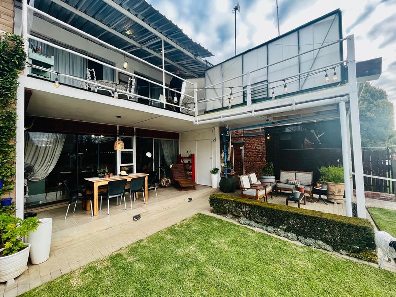 A spacious rear exterior featuring a covered patio area, an upper-level balcony, and a well-maintained garden with brick and paved elements.