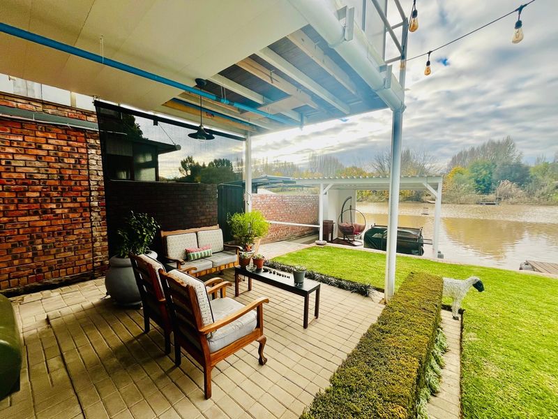 A covered outdoor patio area featuring paved flooring and exposed brickwork, overlooking a landscaped garden and waterfront.