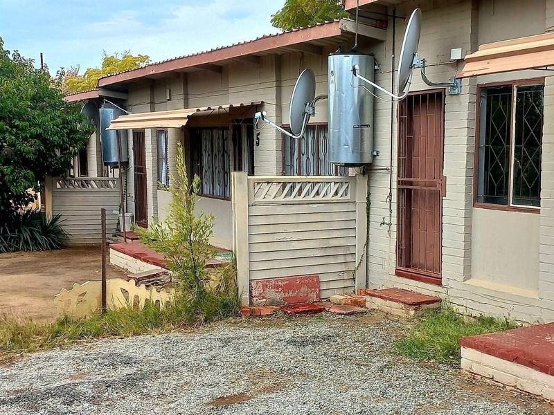 Front elevation of a cluster unit featuring external geysers, security gates, and basic brick construction with a gravel driveway approach.