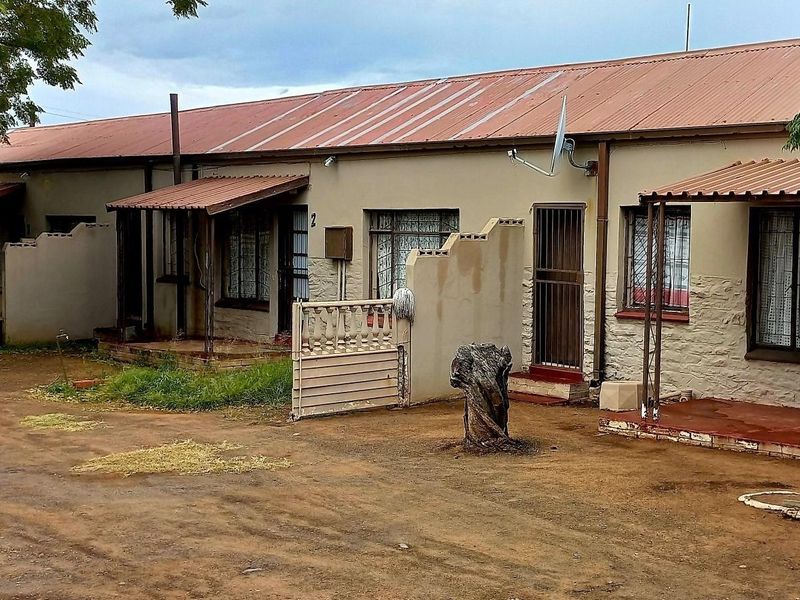 Front elevation of a cluster property featuring a corrugated metal roof, security gates, and individual patio entrances with unpaved ground access.