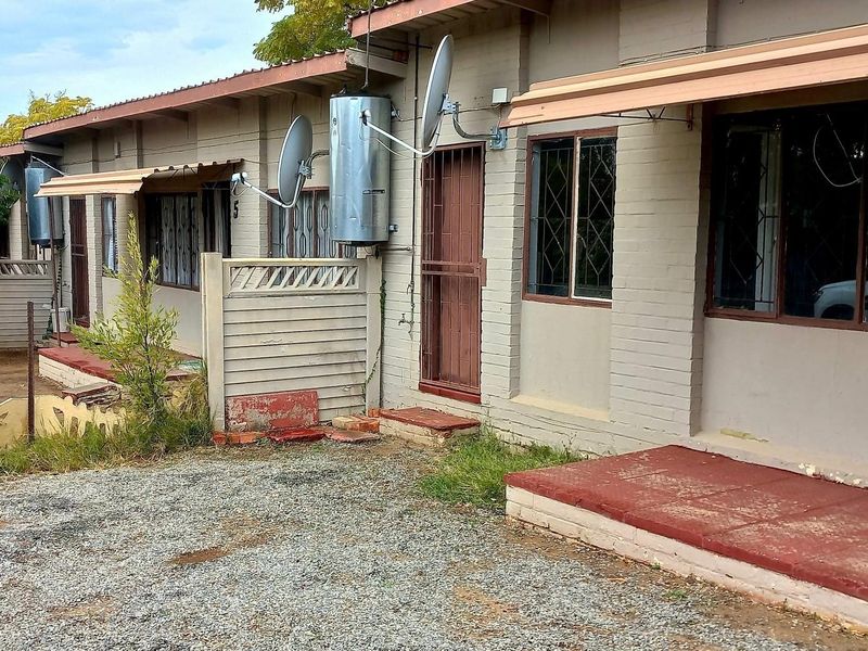 Front exterior view of a cluster unit featuring brick facade, security gates, and external geyser installations with a gravel driveway area.