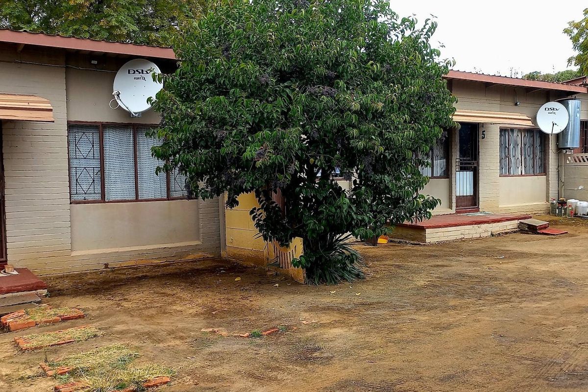 Front elevation of a cluster property featuring brick walls, metal roofing, and security gates, with an unpaved driveway area.