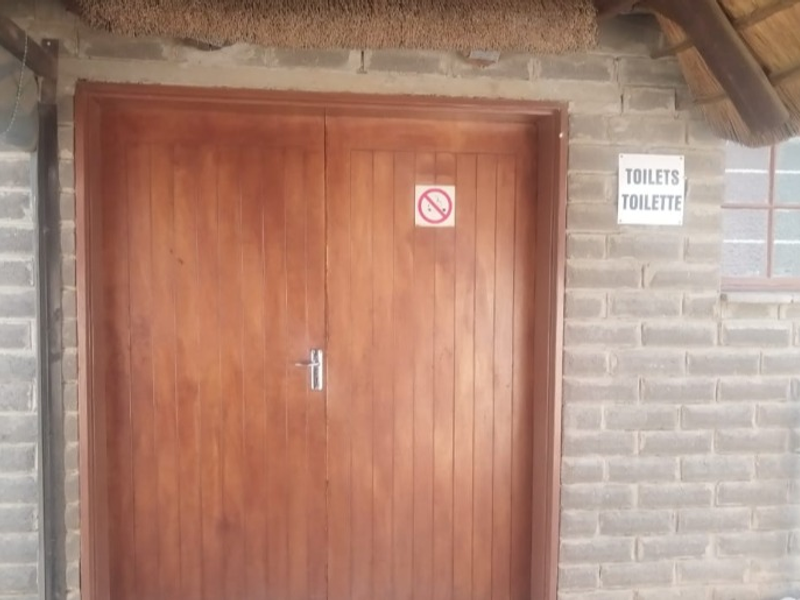 Exterior view of a lodge facility featuring exposed brick walls, a thatched roof overhang, and timber double doors leading to the toilets.