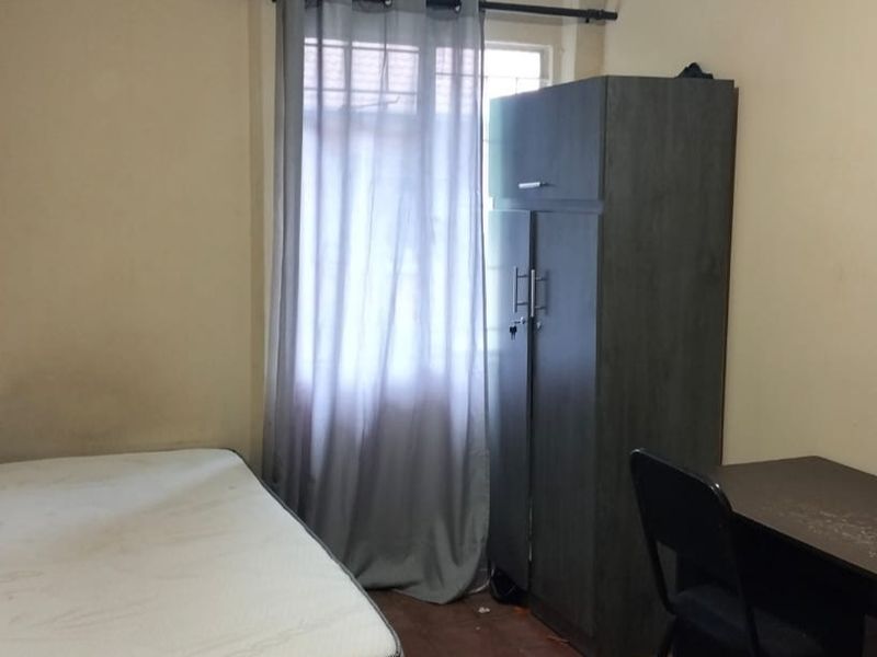 A compact bedroom featuring a window with curtain rod, neutral wall finishes, and basic flooring, suitable for a single occupant.