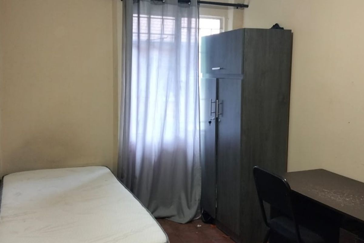 A compact bedroom featuring a window with curtain rod, neutral wall finishes, and basic flooring, suitable for a single occupant.