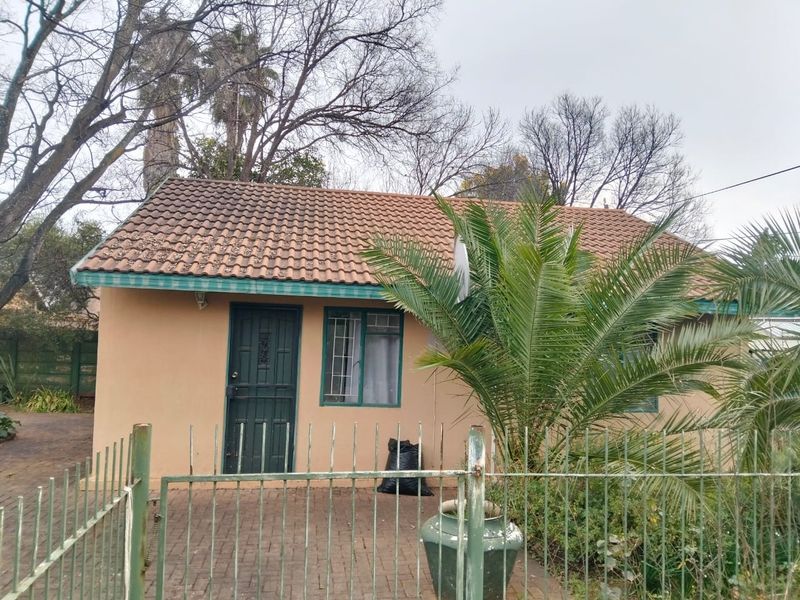 Front elevation of a single-story house featuring a tiled roof, security gate, and paved driveway area, showing signs of exterior wear.