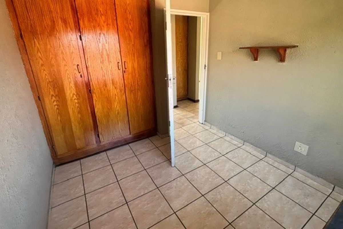 A compact bedroom featuring built-in wooden wardrobes, ceramic tiled flooring, and a wall-mounted shelf, offering a functional space ready for modernization.