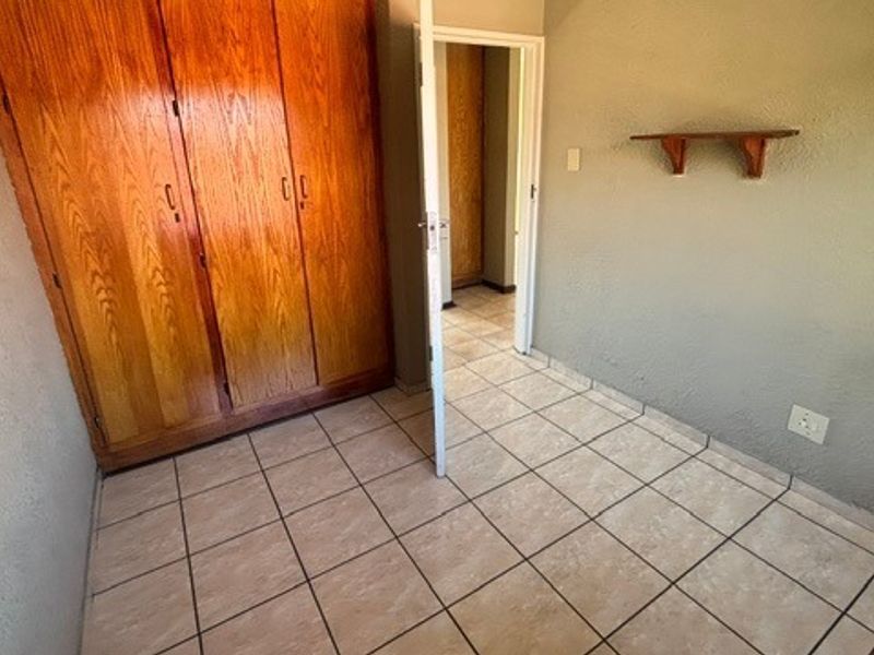 A compact bedroom featuring built-in wooden wardrobes, ceramic tiled flooring, and a wall-mounted shelf, offering a functional space ready for modernization.