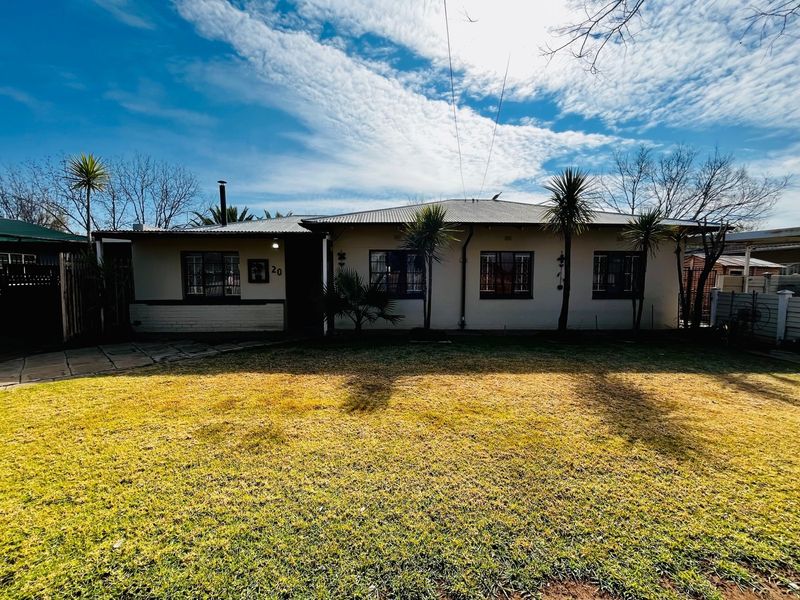 Front elevation of a single-story house featuring a corrugated metal roof, security bars on windows, and a paved driveway with a front lawn.