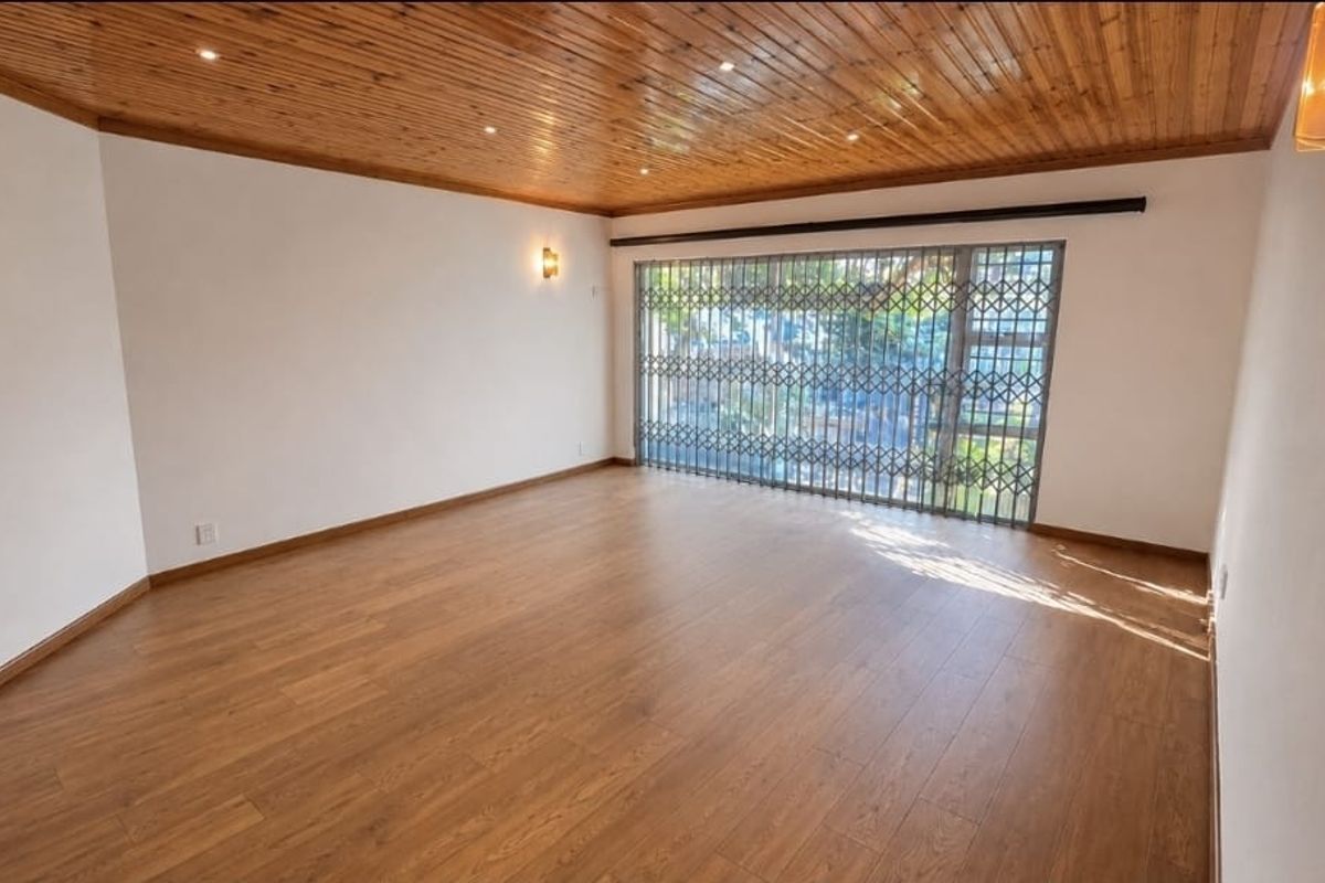 Spacious living area featuring warm laminate flooring, a distinctive timber-clad ceiling with recessed lighting, and a large window secured by a metal gate.