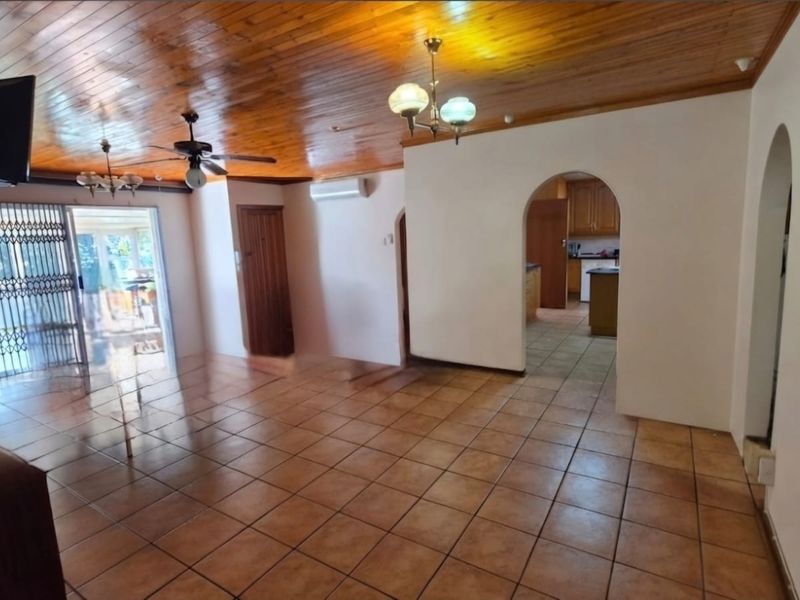 Spacious open-plan living area featuring timber tongue and groove ceilings, ceramic floor tiles, and an arched entryway leading to the kitchen.