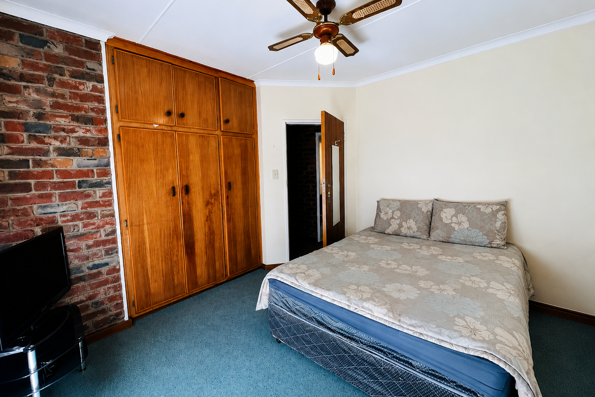 A bedroom featuring an exposed brick wall, built-in wooden wardrobes, and a ceiling fan, offering a functional space with potential for modern updates.