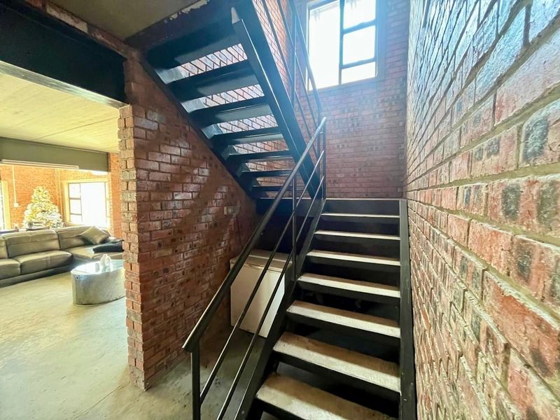 Industrial-style hallway featuring exposed brick walls, a metal staircase with matching handrails, and concrete flooring, leading into an open-plan living area.