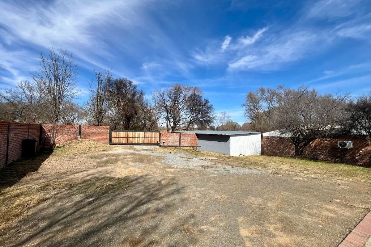 A spacious rear yard featuring a brick boundary wall, a sliding gate, an outbuilding, and an asphalt driveway area.