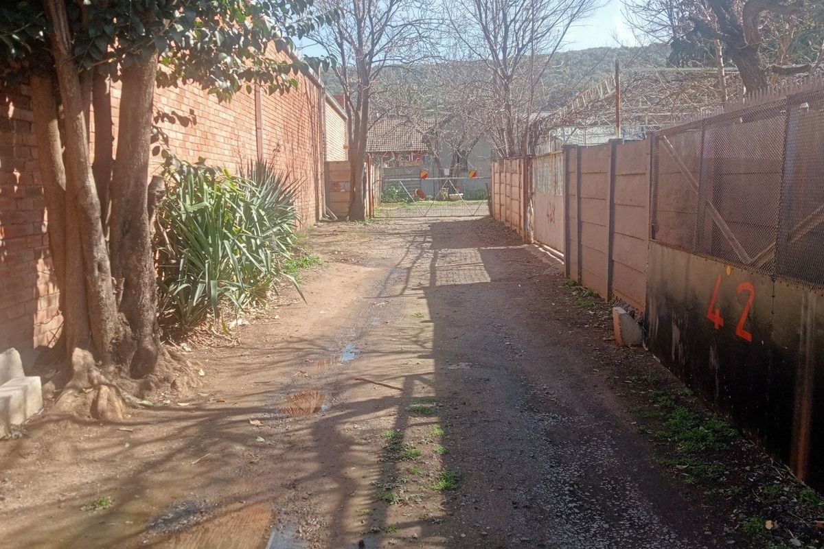 A narrow rear access path or alleyway featuring brick and precast concrete boundary walls with metal security fencing.
