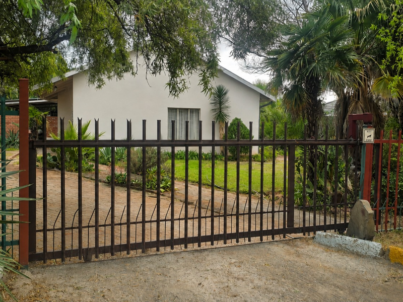 Front view of the property featuring a metal sliding gate, paved driveway, and established garden area.