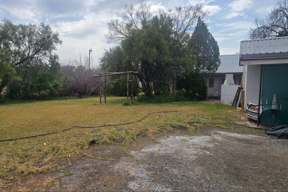 A view of the rear garden area featuring a lawn, mature trees, a pergola structure, and a portion of the building with a metal roof.