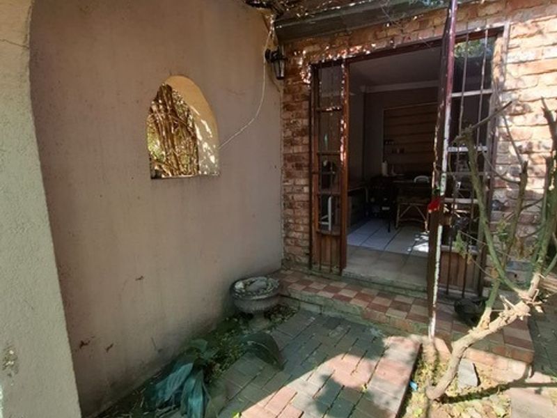 A small, weathered outdoor patio area featuring brick paving, an arched wall niche, and a metal security gate leading into the home.