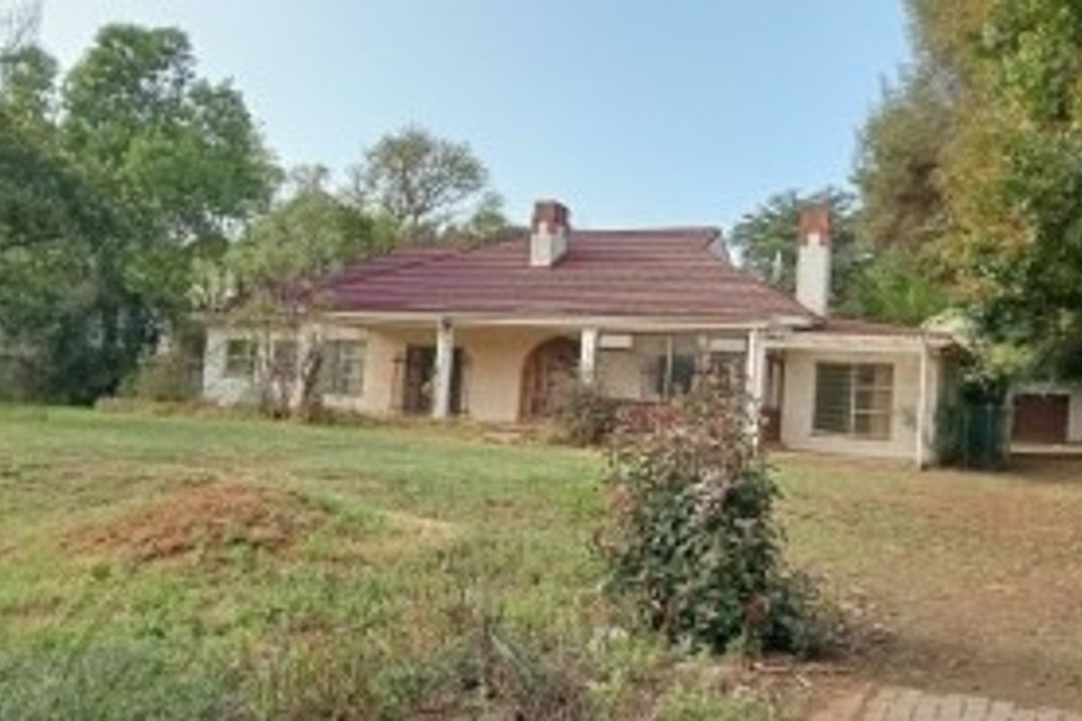 A rear view of a single-story house featuring a tiled roof, chimneys, and a covered patio set within a large, overgrown garden.
