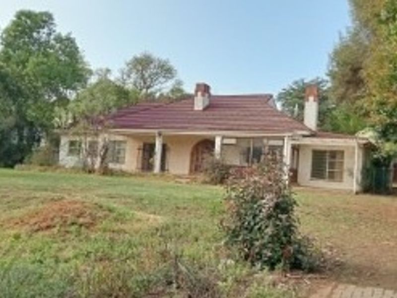 A rear view of a single-story house featuring a tiled roof, chimneys, and a covered patio set within a large, overgrown garden.
