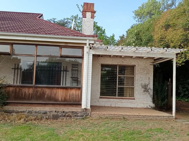 Rear view of the house featuring a covered patio area, brick chimney, and large windows with security bars, set against a mature garden.