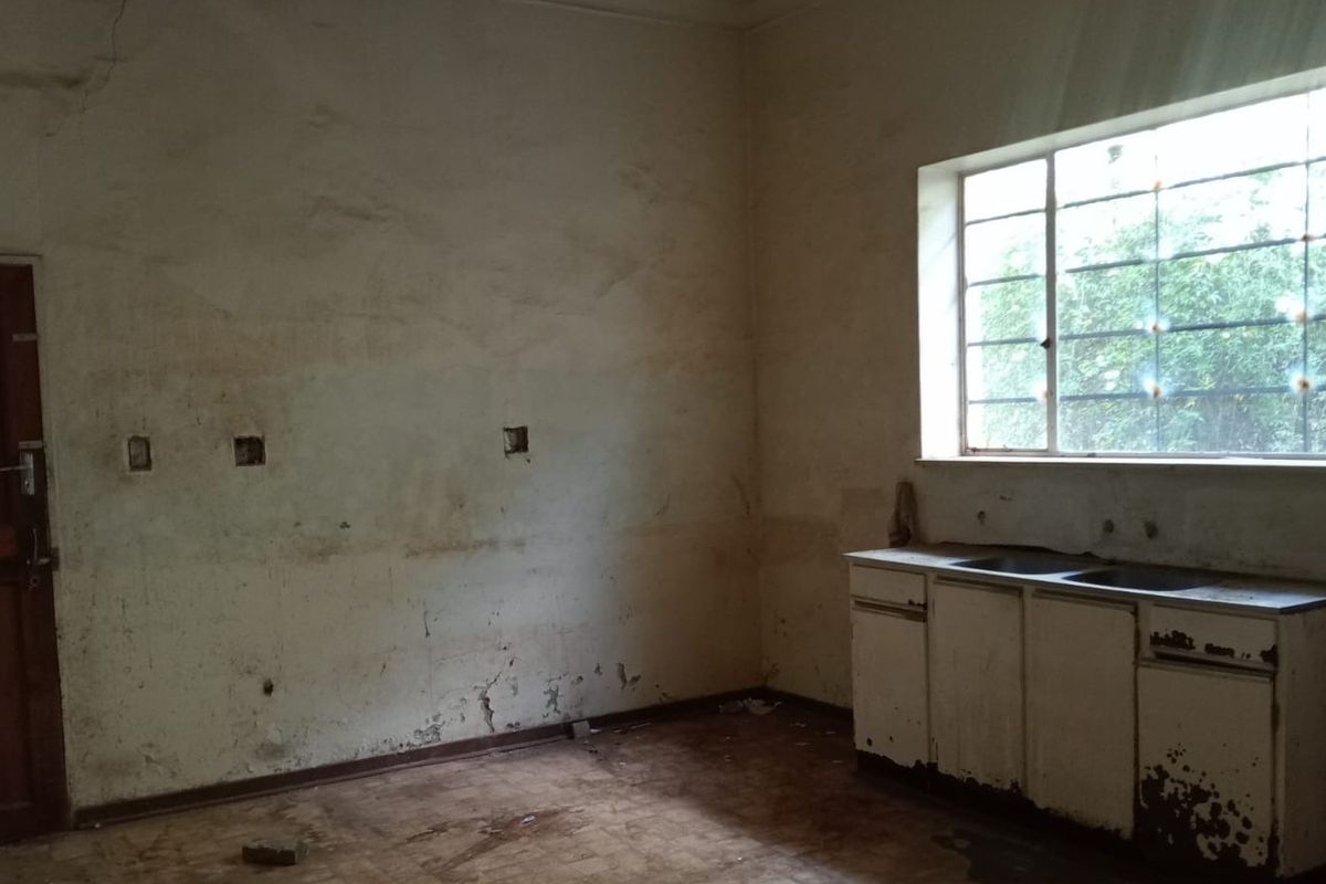 An empty, dilapidated kitchen space featuring original cabinetry, a stainless steel sink, and significant wall surface damage requiring renovation.