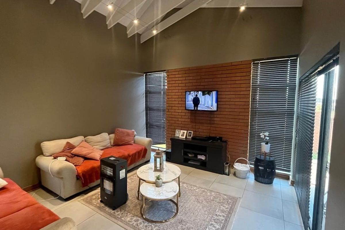 Spacious living room featuring high ceilings with exposed rafters, a prominent exposed brick feature wall, tiled flooring, and large windows with venetian blinds.