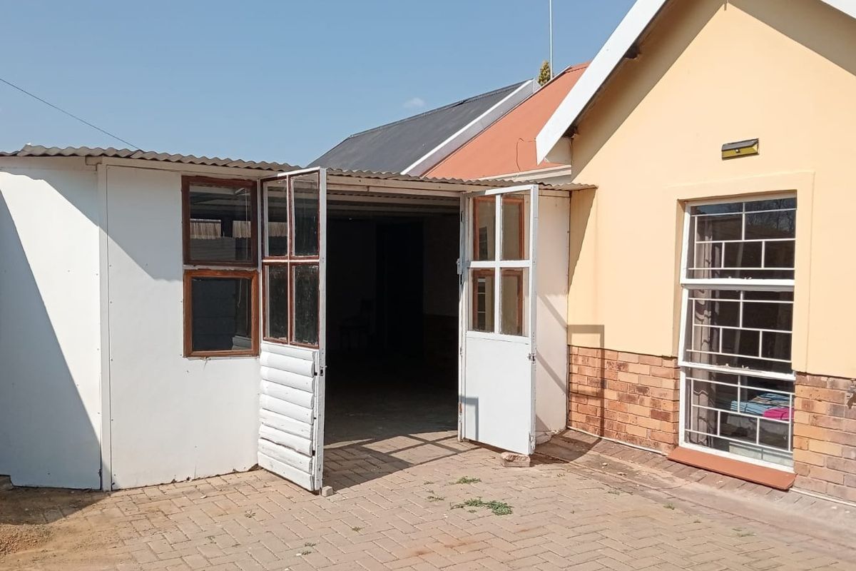 Rear exterior view showing a paved courtyard area, brick facade, and access to a secondary structure with hinged doors and security-barred windows.