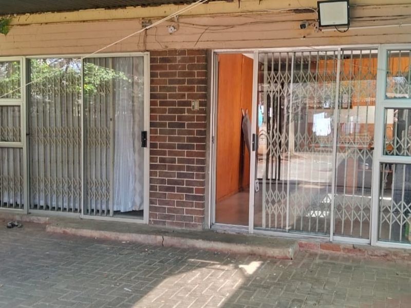A covered patio area featuring brick exterior walls, sliding glass doors with security gates, and paved flooring, showing signs of wear and maintenance needs.