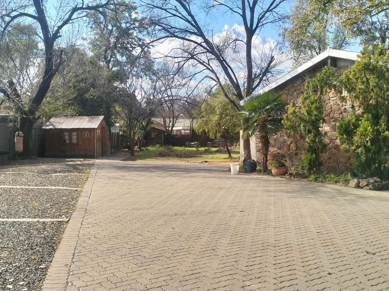 Spacious rear exterior area featuring a paved driveway, gravel parking space, stone-clad building facade, and a detached timber outbuilding set within a garden.