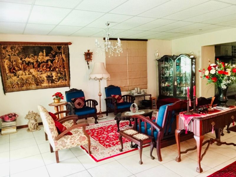 A spacious living room featuring tiled flooring, a drop ceiling, and a central chandelier, offering a traditional layout with ample space for seating.