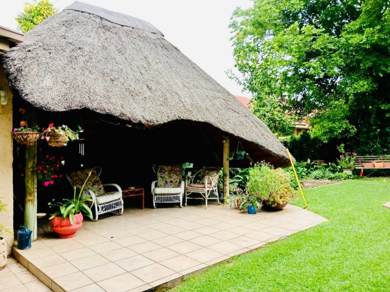 A covered outdoor patio area featuring a traditional thatched roof, tiled flooring, and timber support posts, overlooking a lush green garden.