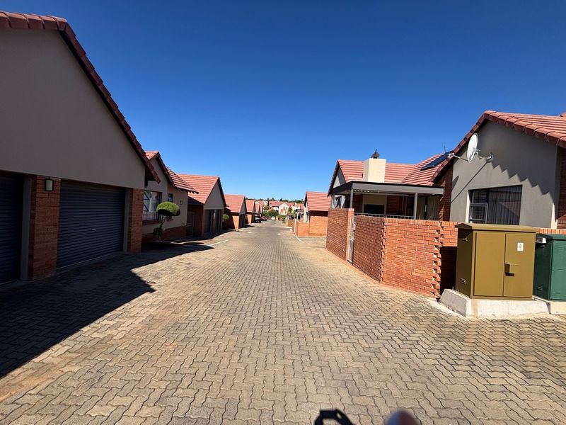 A view of the paved complex driveway lined with residential townhouse units featuring brick walls and tiled roofs under a clear blue sky.