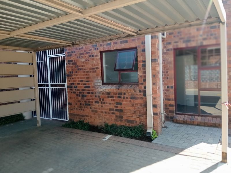 A covered carport area with brick exterior walls, paved flooring, and secure metal gate access to the townhouse entrance.