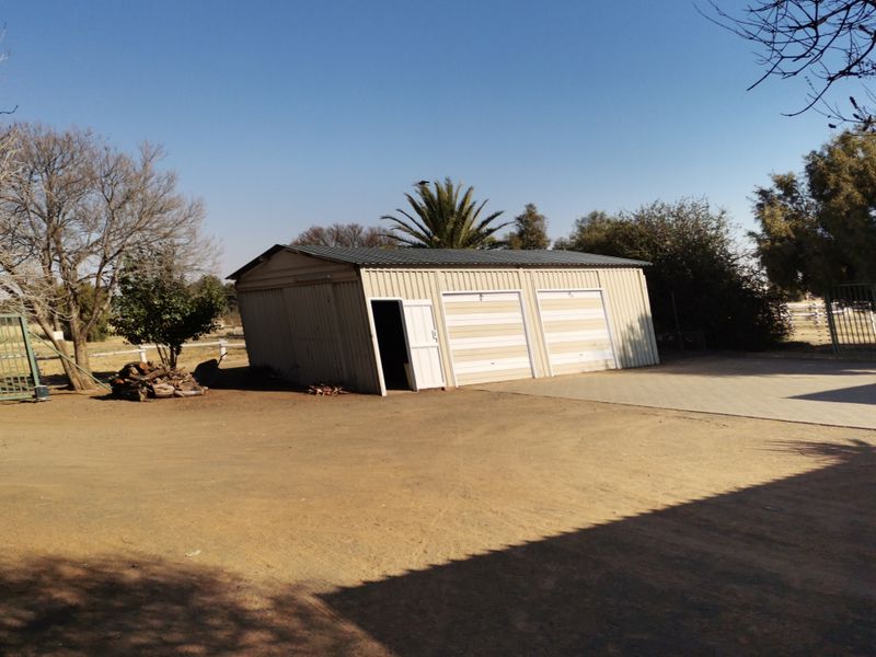 A detached industrial-style metal garage featuring double garage doors and a side entrance, situated on a dirt and concrete driveway.