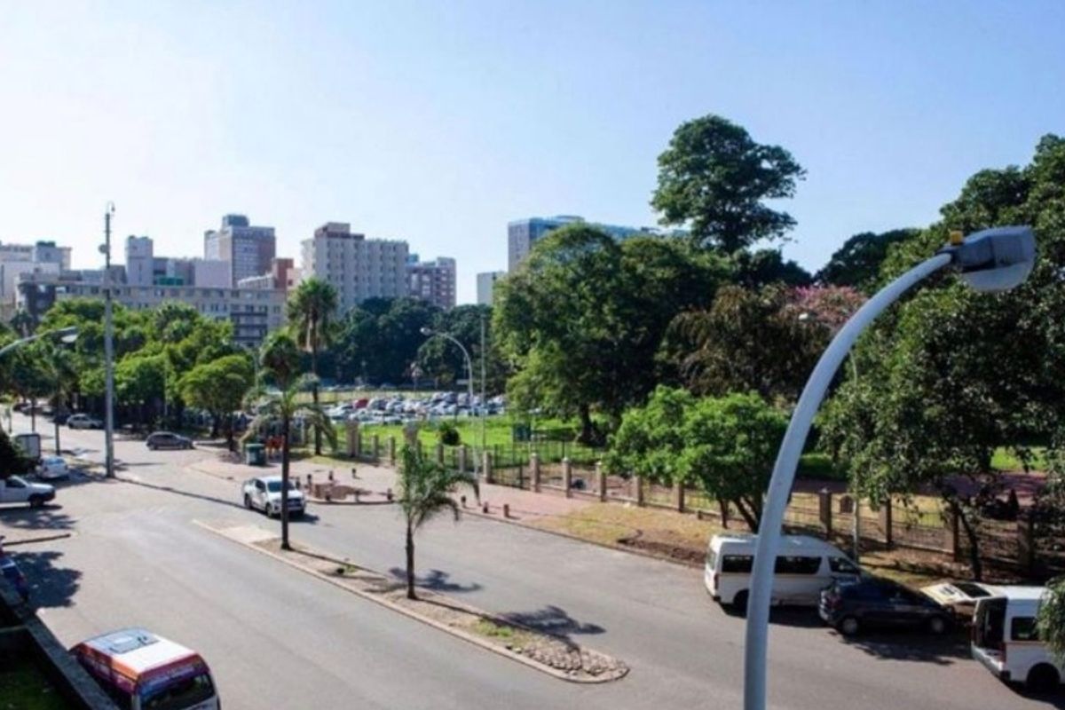 An elevated street view showing the surrounding urban environment, including paved roads, mature trees, and perimeter fencing near the property.