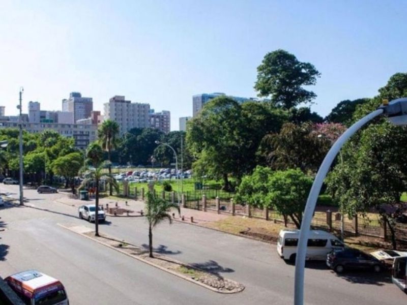 An elevated street view showing the surrounding urban environment, including paved roads, mature trees, and perimeter fencing near the property.